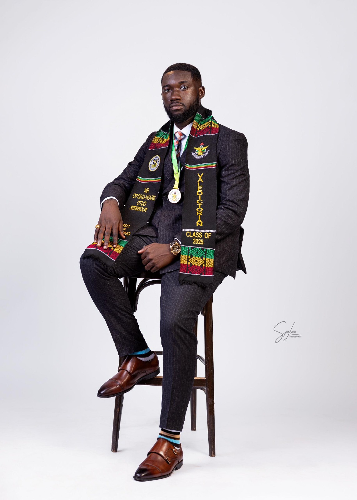 A graduate seated on a stool wearing the Ghana Kente sash, embroidered with his name, two crests and "Valedictorian, Class of 2025".